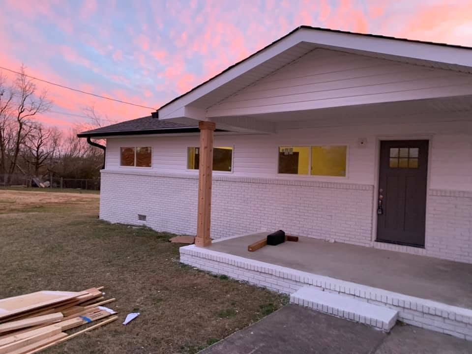 A single-story house with white-painted brick, a covered front porch with a cedar post and a dark gray front door, under a pink sunset sky, with lumber stacked in the yard.