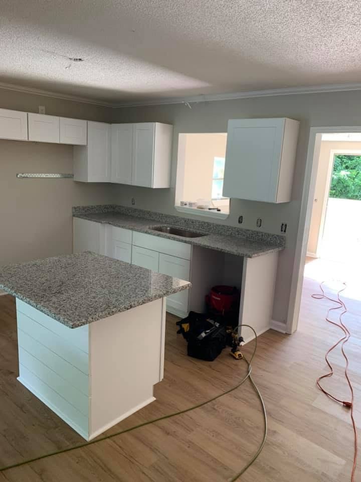 A kitchen being installed: white shaker cabinets, speckled gray countertops, an island with shiplap-style paneling, and tools and cords on the wood-look floor.