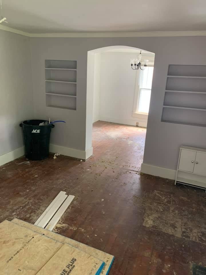 A living room with lavender walls, two built-in shelves and an arched opening to a dining room, with the old wood floor still bare and a trash can in the corner.