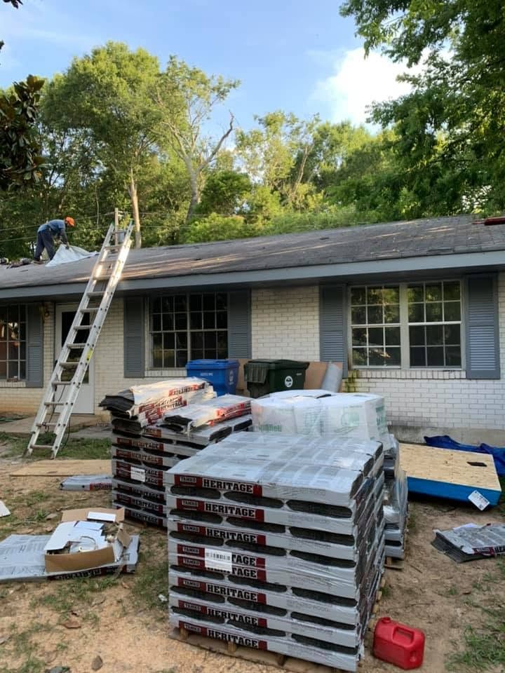 A brick ranch house with a roofer on the roof, a ladder against the eave and pallets of shingles stacked in the yard.