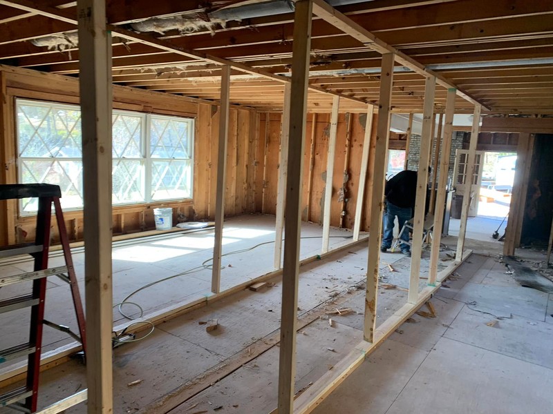 The inside of a house stripped down to the studs and joists, with a new row of framed wall studs in the foreground, large windows on the left and a worker bending over at the back.