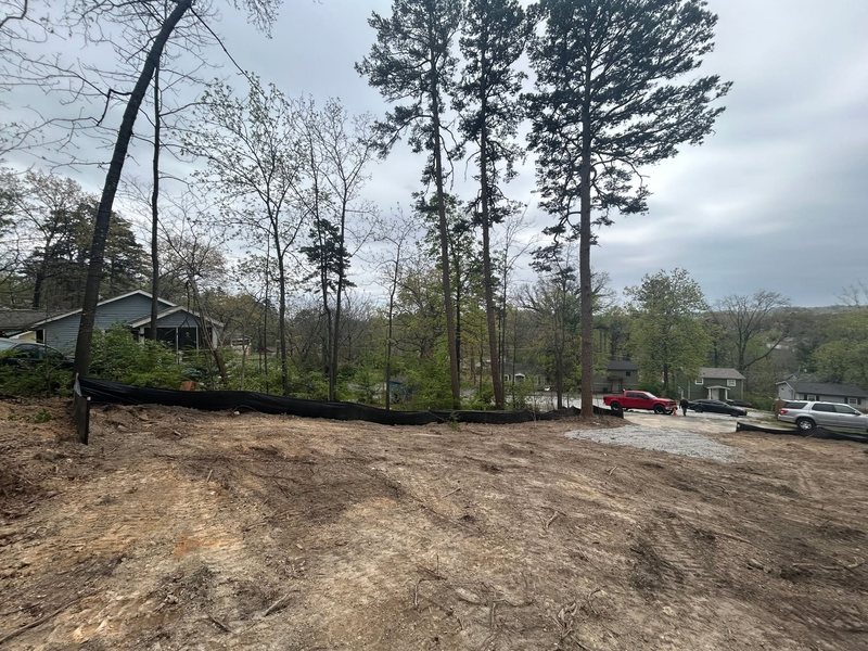 Bare, graded dirt on a hillside lot, with black silt fence along the edge, a gravel entrance on the right and tall pines behind